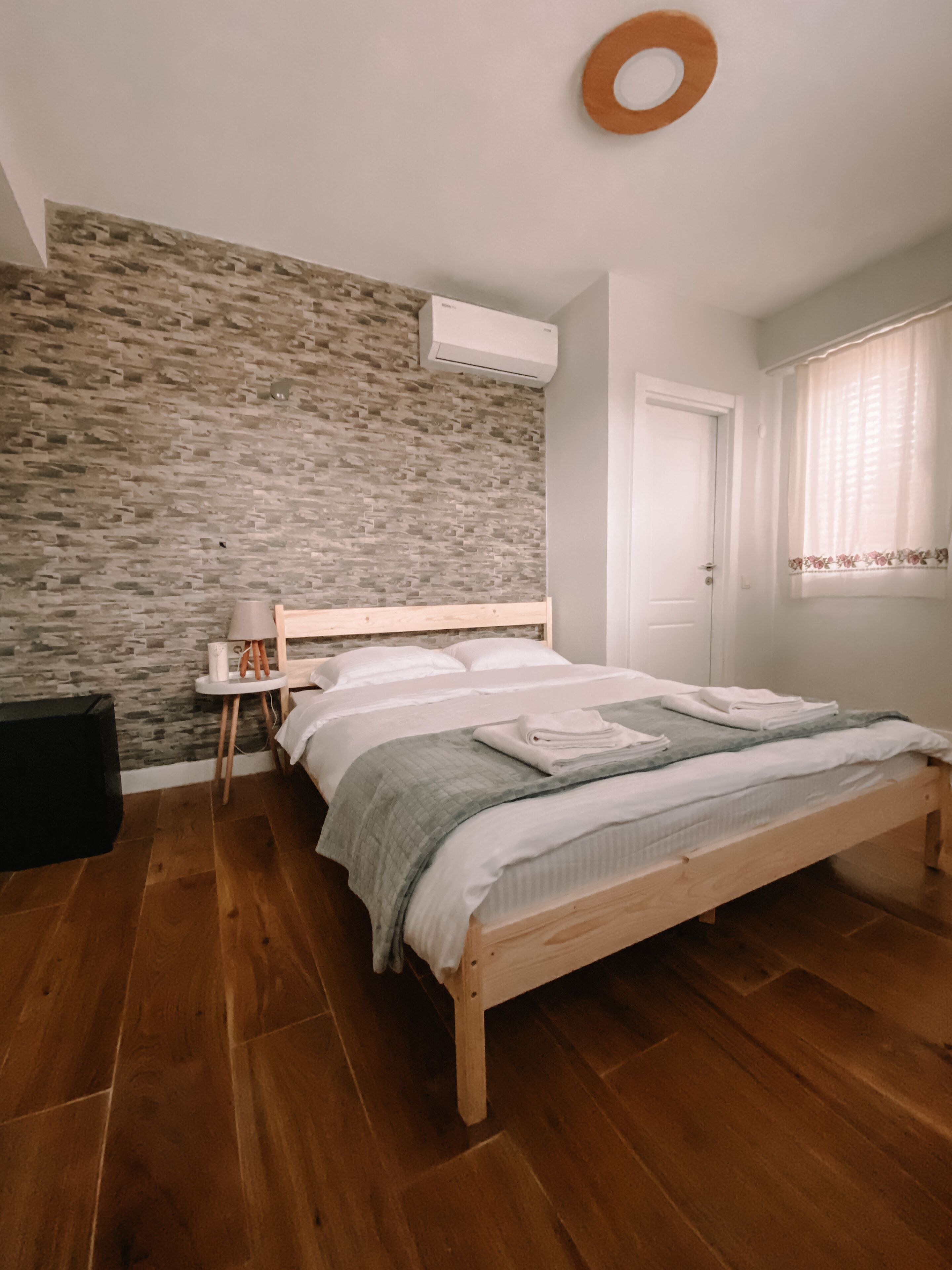 A bedroom with a wooden bed, a small bedside table, a wall-mounted air conditioner, and a window with curtains.