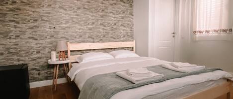 A bedroom with a wooden bed, a small bedside table, a wall-mounted air conditioner, and a window with curtains.