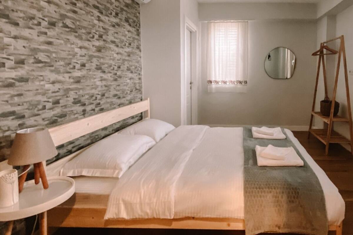A bedroom with a wooden bed, a round mirror, a wooden side table, and a brick wall.