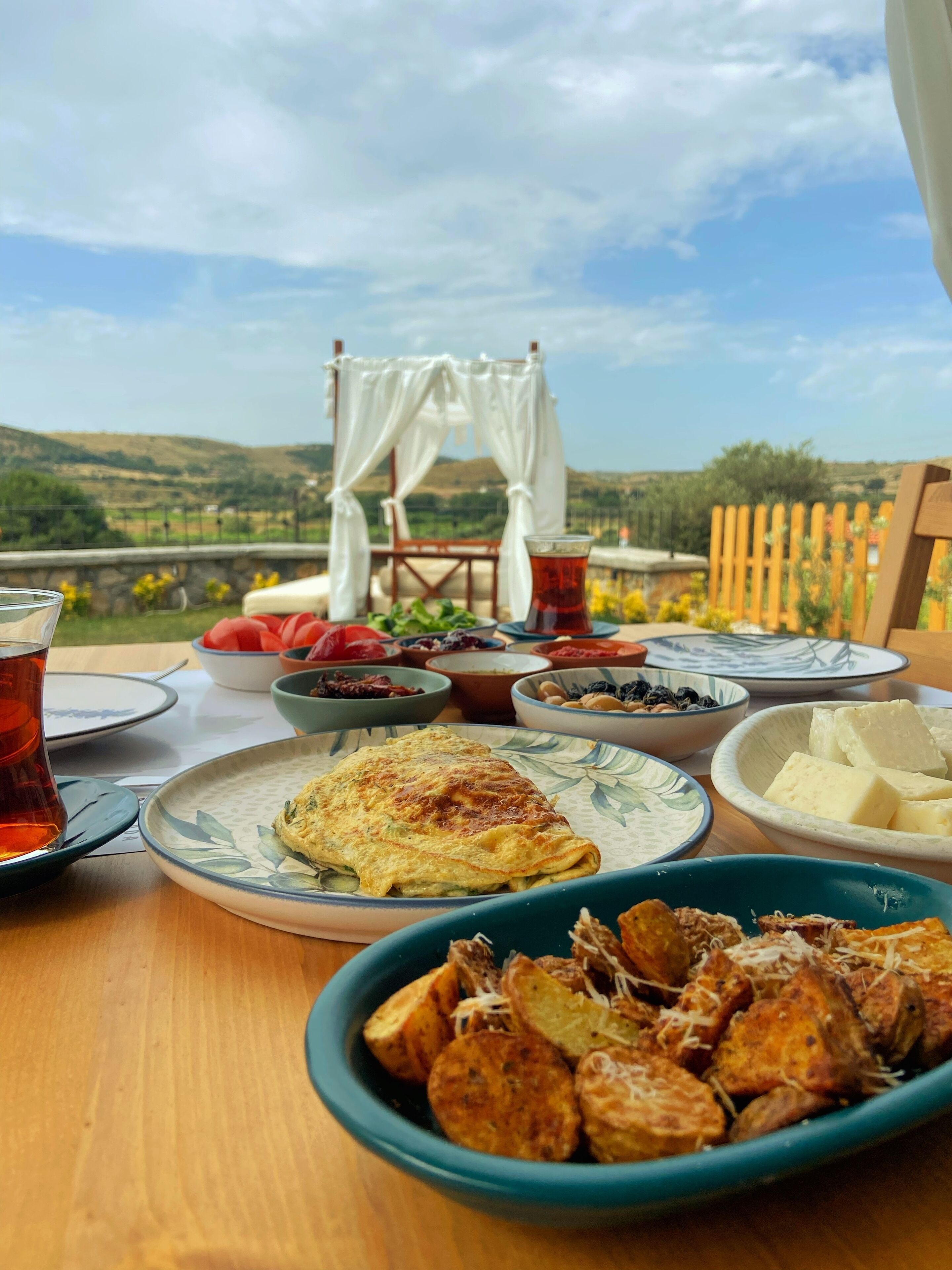 A table set with various dishes, including a plate of roasted potatoes, a bowl of cheese, and a glass of tea, with a scenic view in the background.