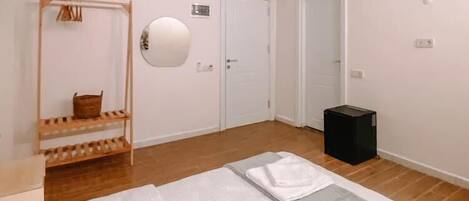 Une chambre à coucher avec un lit, une étagère en bois où sont posées des serviettes, un miroir et une poubelle noire.