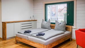 A bedroom with a wooden bed, grey bedding, a red chair, and a window with blinds. - Gästewohnung Storchennest (Teichland)
