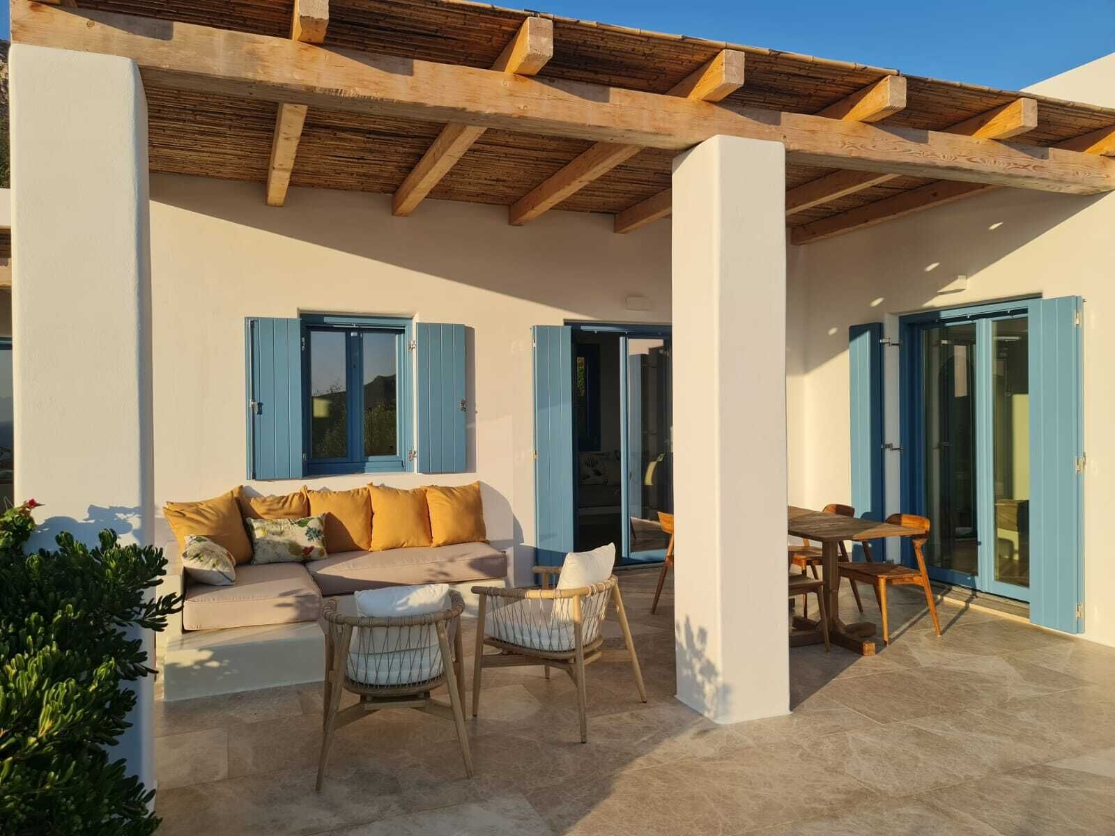 A covered outdoor seating area with a sofa and chairs, a table, and a window with blue shutters.