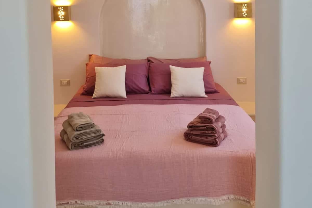 Un dormitorio con cama, almohadas y cabecera. Hay dos luces fijadas a la pared a ambos lados de la cama.