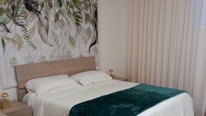 Un lit bien fait, avec du linge de lit blanc et un plaid turquoise, une tête de lit en bois, une table de chevet avec une lampe, et un mur orné d’un motif de feuilles.