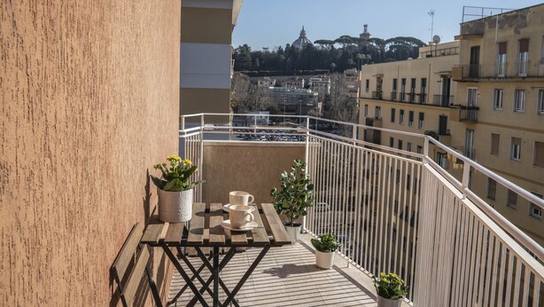 A balcony with a wooden table, two potted plants, and a view of a cityscape with buildings and a dome in the distance. - Vintage Apartment Cipro (Rome)