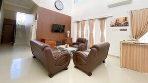 Amplia sala de estar con techo alto, televisor de pantalla plana montado en una pared de paneles de madera, un cómodo sofá marrón, una mesa de centro de vidrio y un comedor con mesa y sillas de madera.