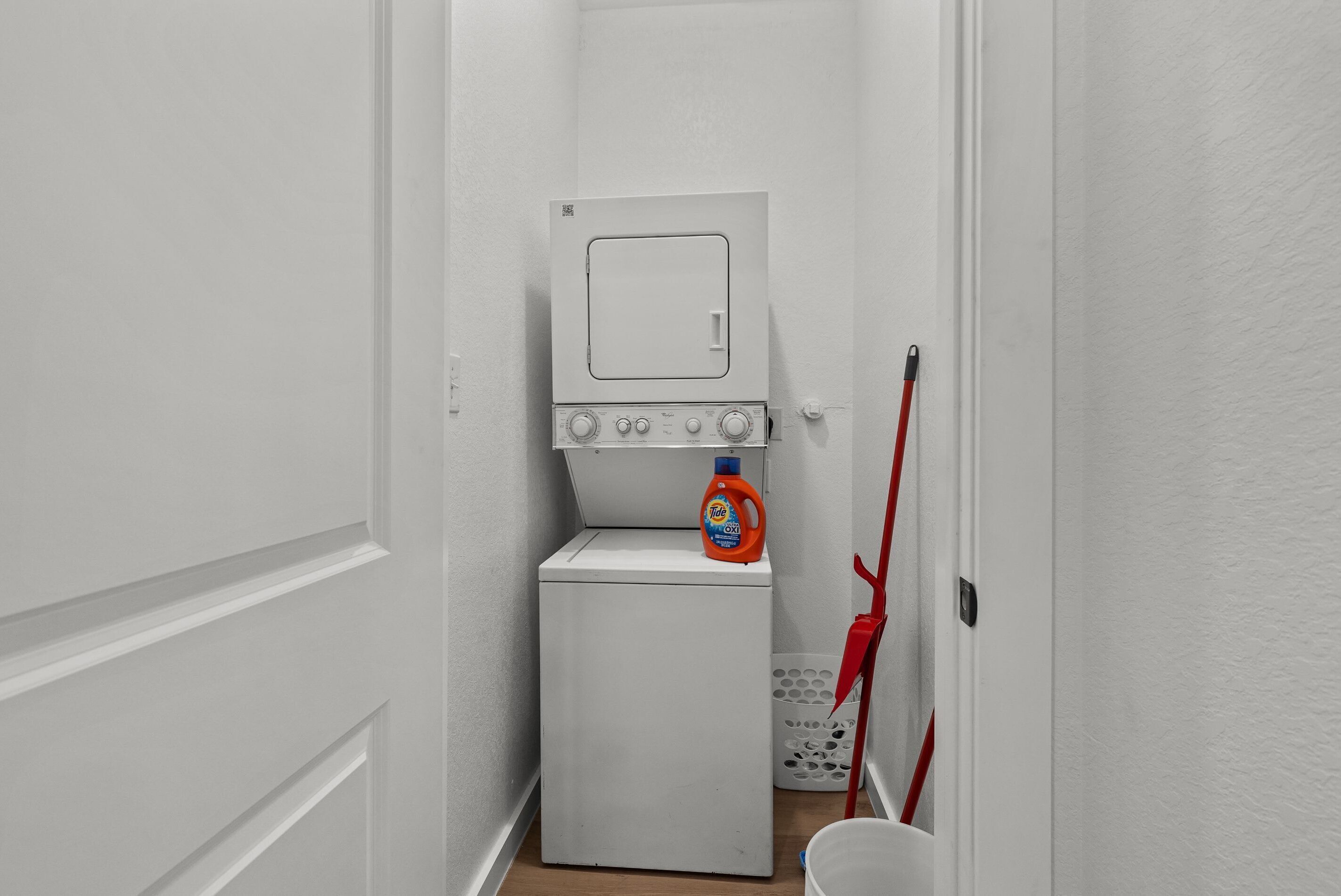 A laundry room with a washing machine and dryer, a laundry detergent bottle, and a red mop leaning against the wall.