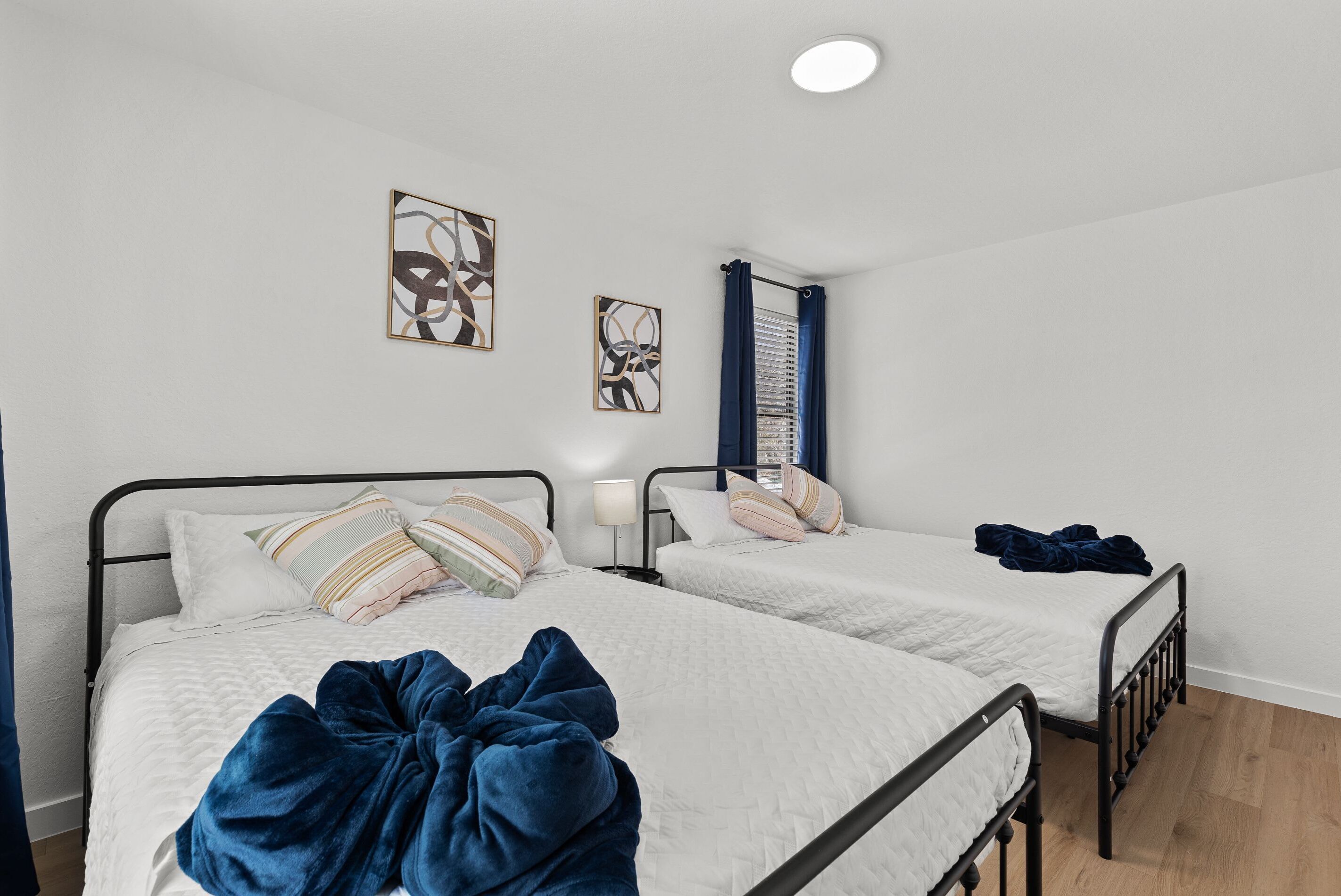 A bedroom with two single beds, each with a white quilted mattress and striped pillows. There are abstract wall art pieces above the beds, and a window with blue curtains.