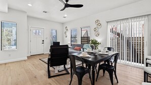 Un espace repas moderne avec une table et des chaises noires, un ventilateur de plafond et une vue sur l’extérieur grâce à de grandes fenêtres.