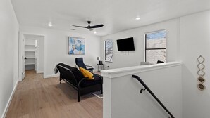 Un salon moderne avec un canapé noir, un ventilateur de plafond, un téléviseur à écran plat et une fenêtre équipée de stores.