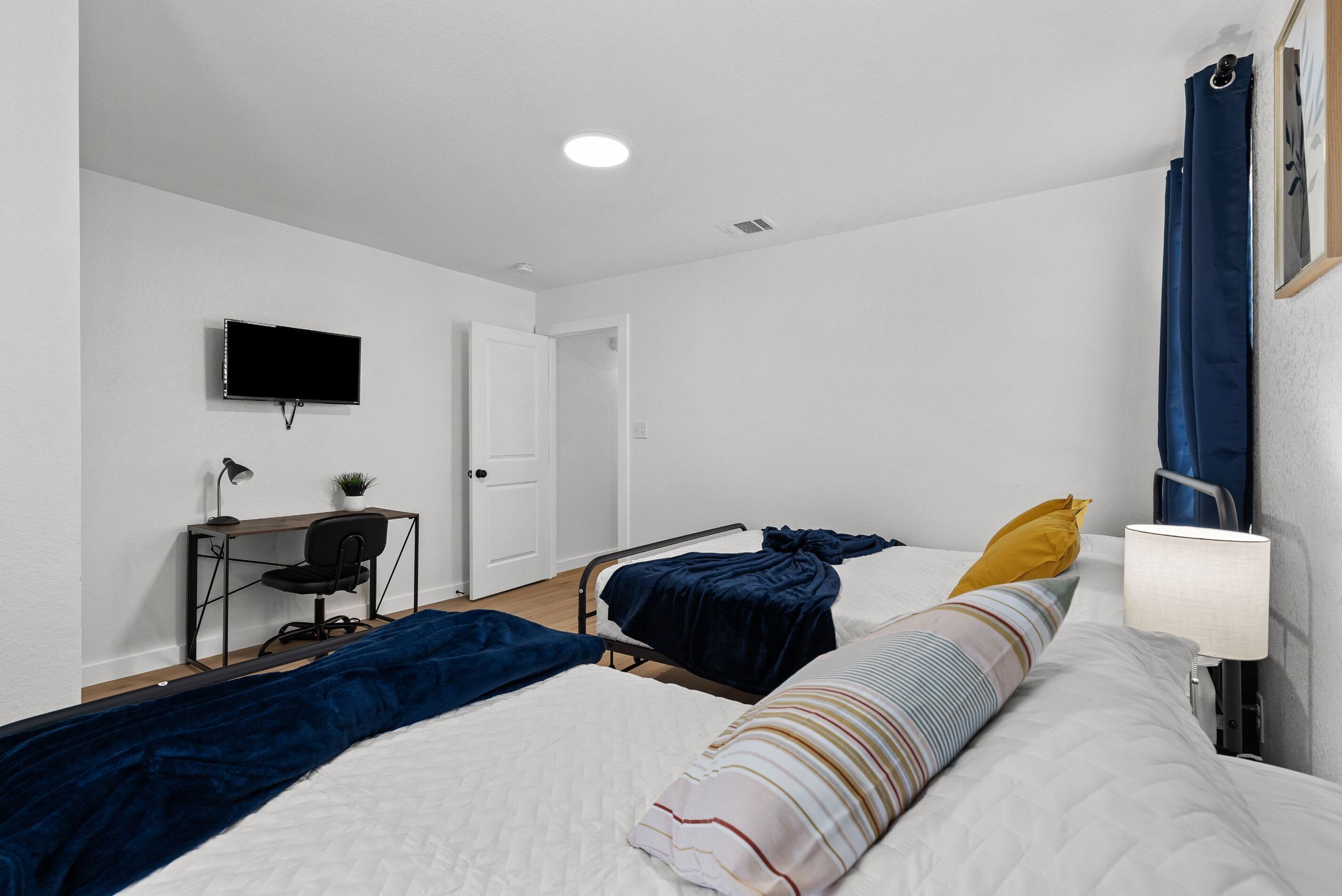 A hotel room with two beds, a desk with a chair, a TV mounted on the wall, and a window with curtains.