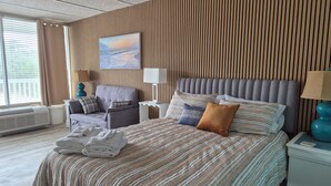 A hotel room with a large bed, a grey upholstered sofa, a wooden slatted wall, and a window with curtains. - Cute Lil' Condo on Intracoastal waterway with beach access and large pool (Atlantic Beach)