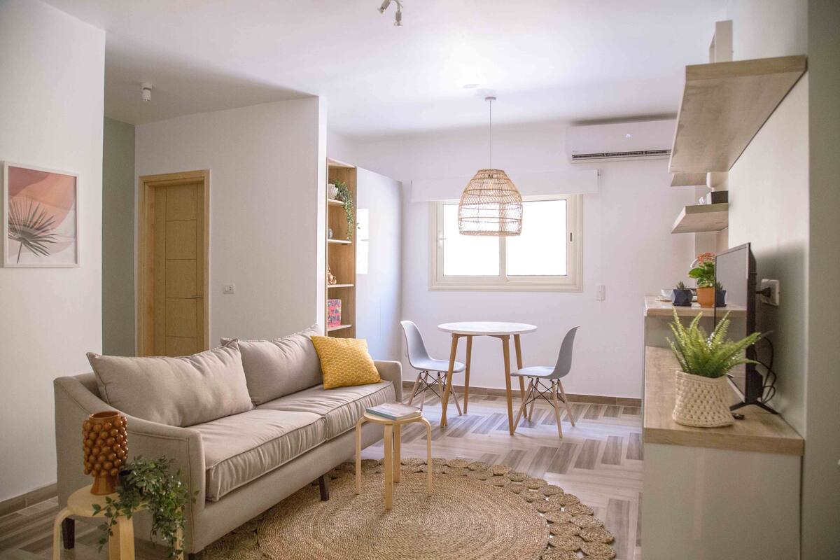 Un salon comprenant un canapé, une petite table et un coin repas avec des chaises. Une fenêtre laisse entrer la lumière naturelle et un luminaire suspendu éclaire la pièce.