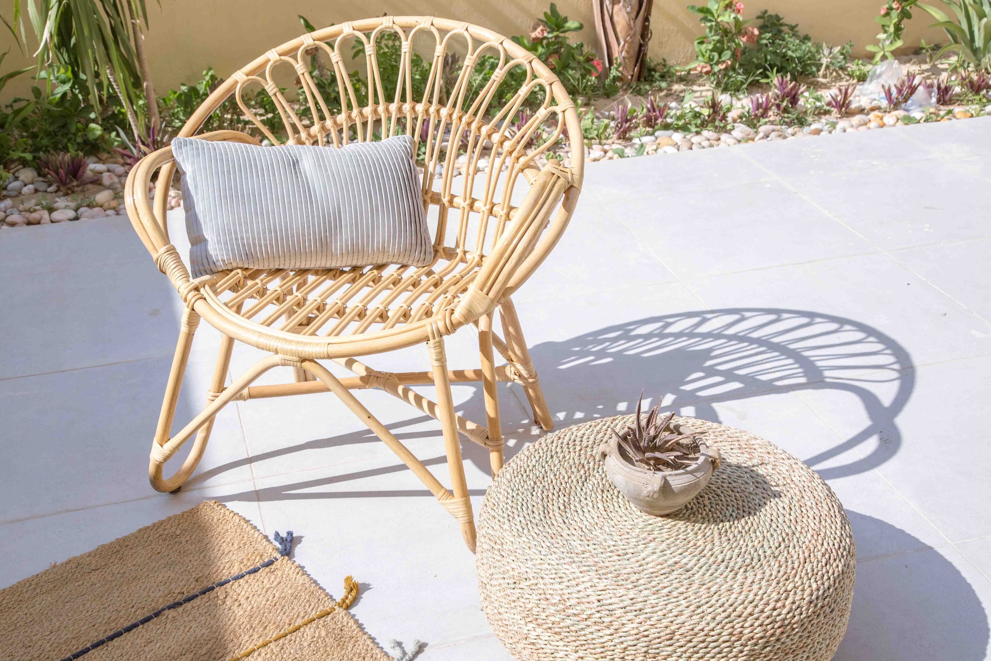 Una silla de mimbre con cojín, un taburete tejido y una pequeña planta en maceta sobre un patio empedrado.