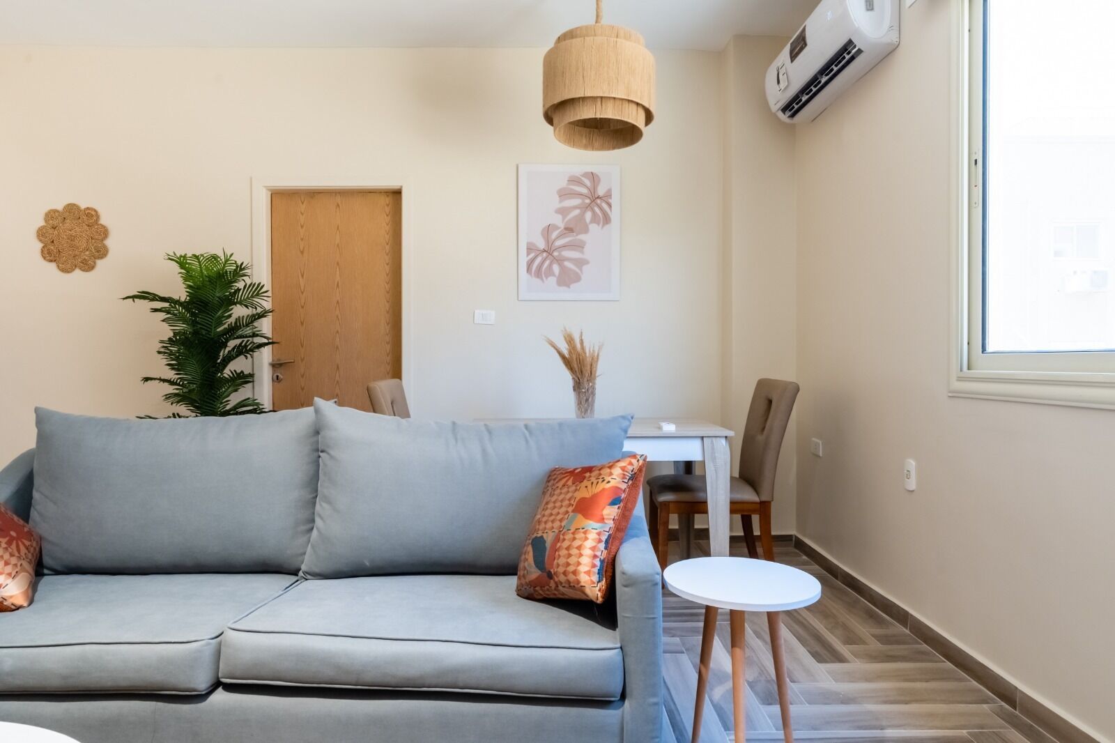 A modern living room with a grey sofa, a small round table, a dining area with a white table and chairs, a wooden door, and an air conditioning unit on the wall.