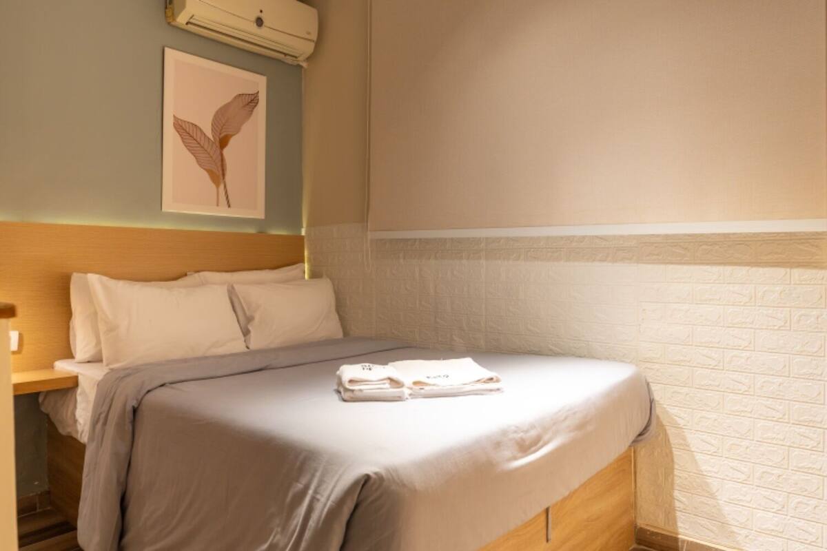 Un lit bien fait, avec une couverture grise et des serviettes pliées, une tête de lit en bois, un climatiseur fixé au mur et une œuvre d’art encadrée au-dessus du lit.