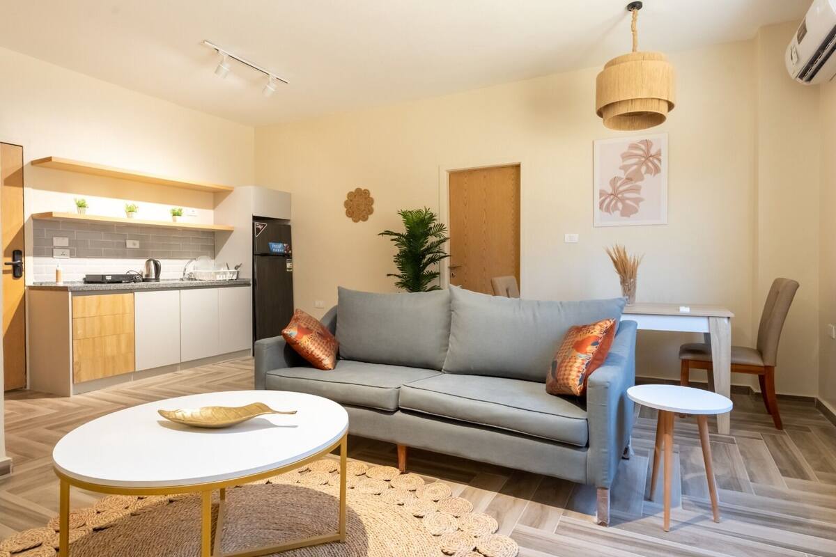 Un espace de vie compact comprenant un coin cuisine, un canapé gris, une table basse ronde et un coin repas avec une petite table et des chaises.