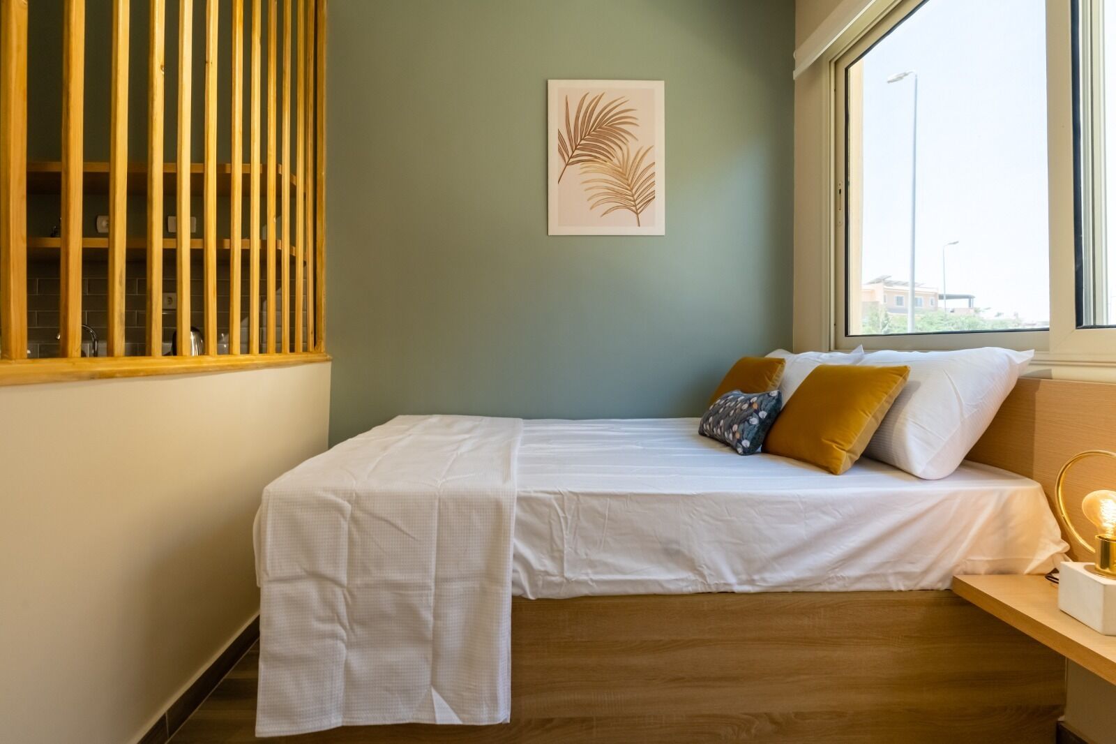 Un lit simple avec une literie blanche, une tête de lit en bois et une fenêtre donnant sur des immeubles.