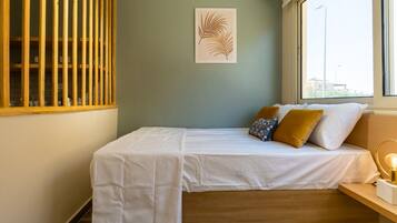 Un lit simple avec une literie blanche, une tête de lit en bois et une fenêtre donnant sur des immeubles.