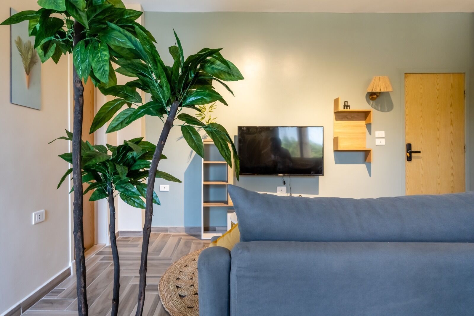 Un salon moderne avec un canapé gris, un téléviseur à écran plat et deux palmiers artificiels.