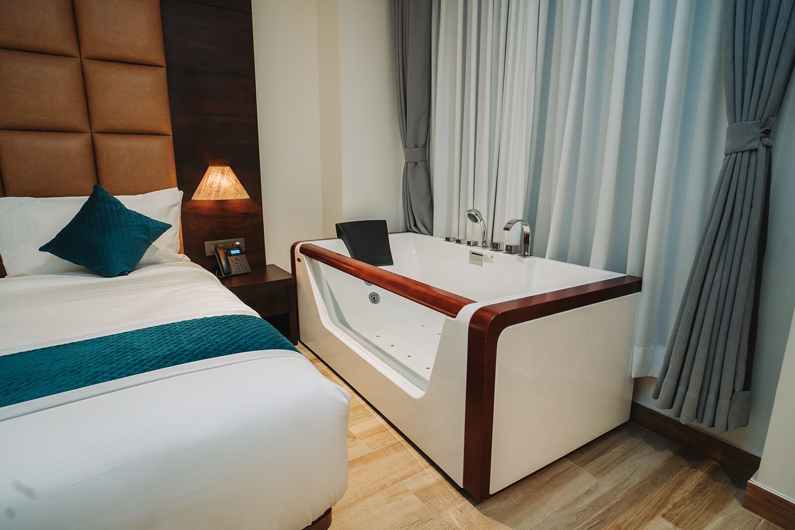 A hotel room with a bed, a bedside table with a lamp, a bathroom with a bathtub, and curtains.