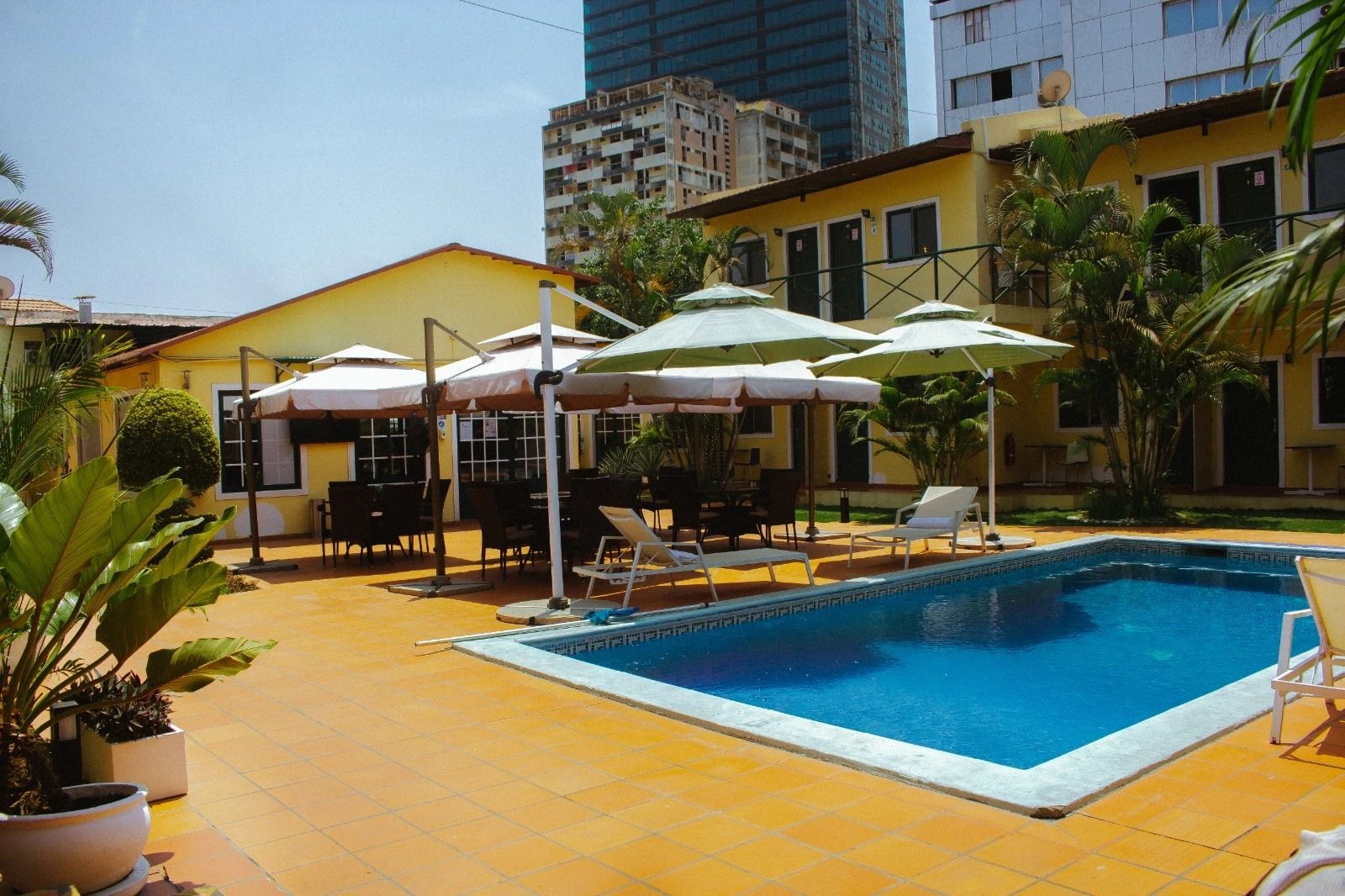 A swimming pool with lounge chairs and umbrellas, surrounded by a yellow building and palm trees.