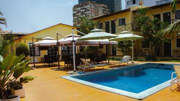 A swimming pool with lounge chairs and umbrellas, surrounded by a yellow building and palm trees.