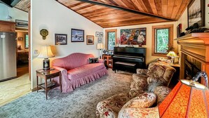 A cozy living room with a wooden ceiling, a piano, a sofa, and a patterned armchair.
