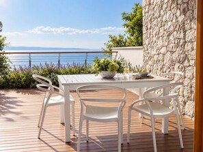 Un espace repas comprenant une table et des chaises, avec vue sur la mer et un mur en pierre.