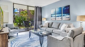 A modern living room with a grey sofa, a glass coffee table, and a view of a tropical garden through a sliding glass door. - NEWLY Updated, Beachfront, WALK out to Beach, Pool, Hot Tub, BBQ! (Kihei)