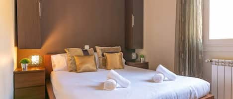 A double bed with white linens, two pillows, a nightstand with a lamp, and a window with curtains.