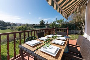Un'area pranzo all'aperto con un tavolo imbandito per quattro persone, circondato da sedie in vimini, su un balcone con vista su un paesaggio verde e edifici in lontananza.