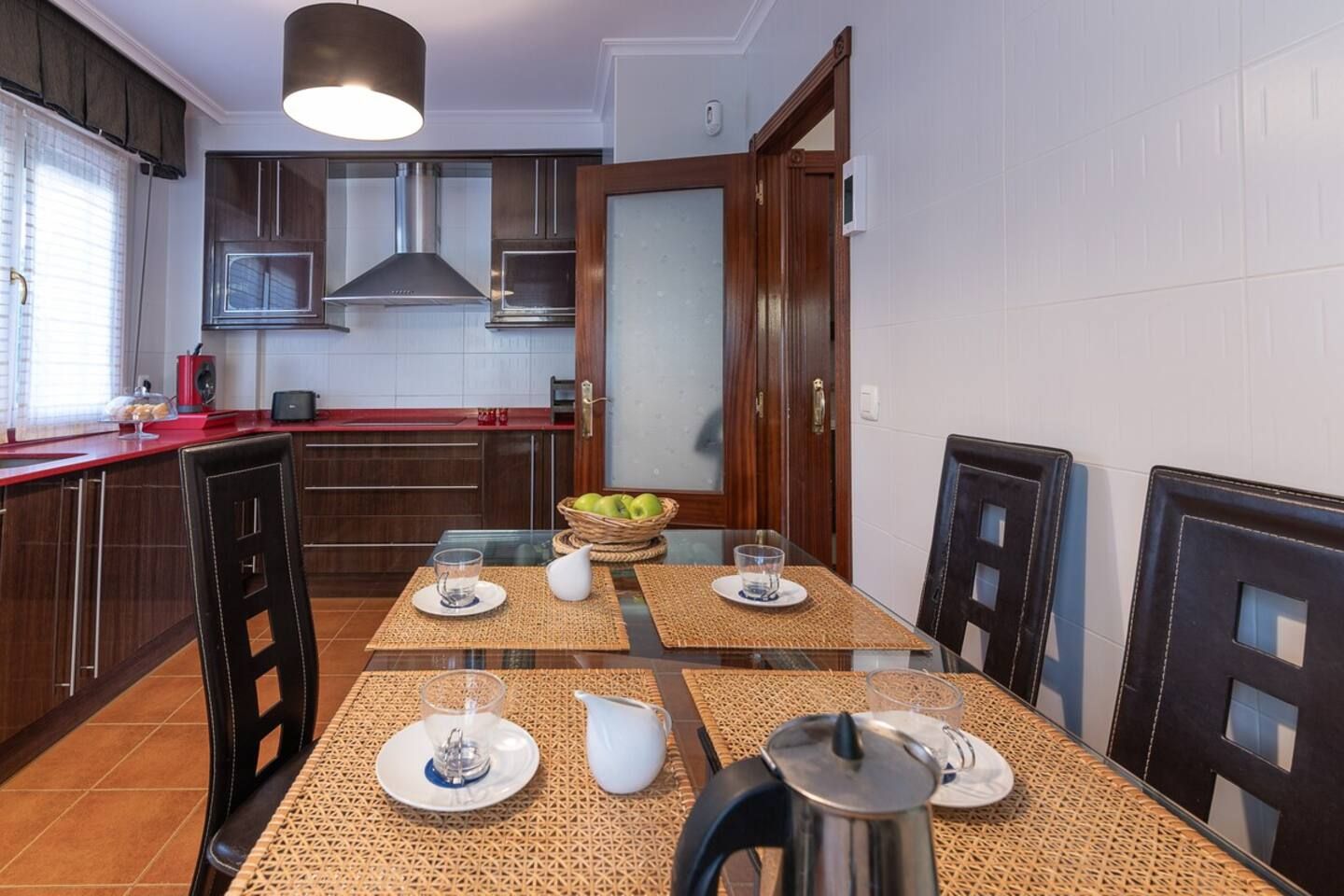 Una cucina moderna con un tavolo da pranzo apparecchiato per quattro persone, dotata di bollitore, bicchieri e una ciotola di frutta.