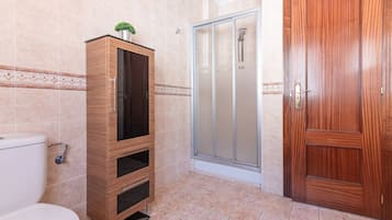 Un baño con inodoro, un armario de madera, cabina de ducha con mampara de vidrio y puerta de madera.
