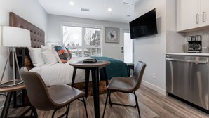 A modern hotel room with a bed, a desk, two chairs, a TV, and a kitchenette. - Sky Nook - Micro Studio - Pool, Hot Tub & Parking (Canmore)