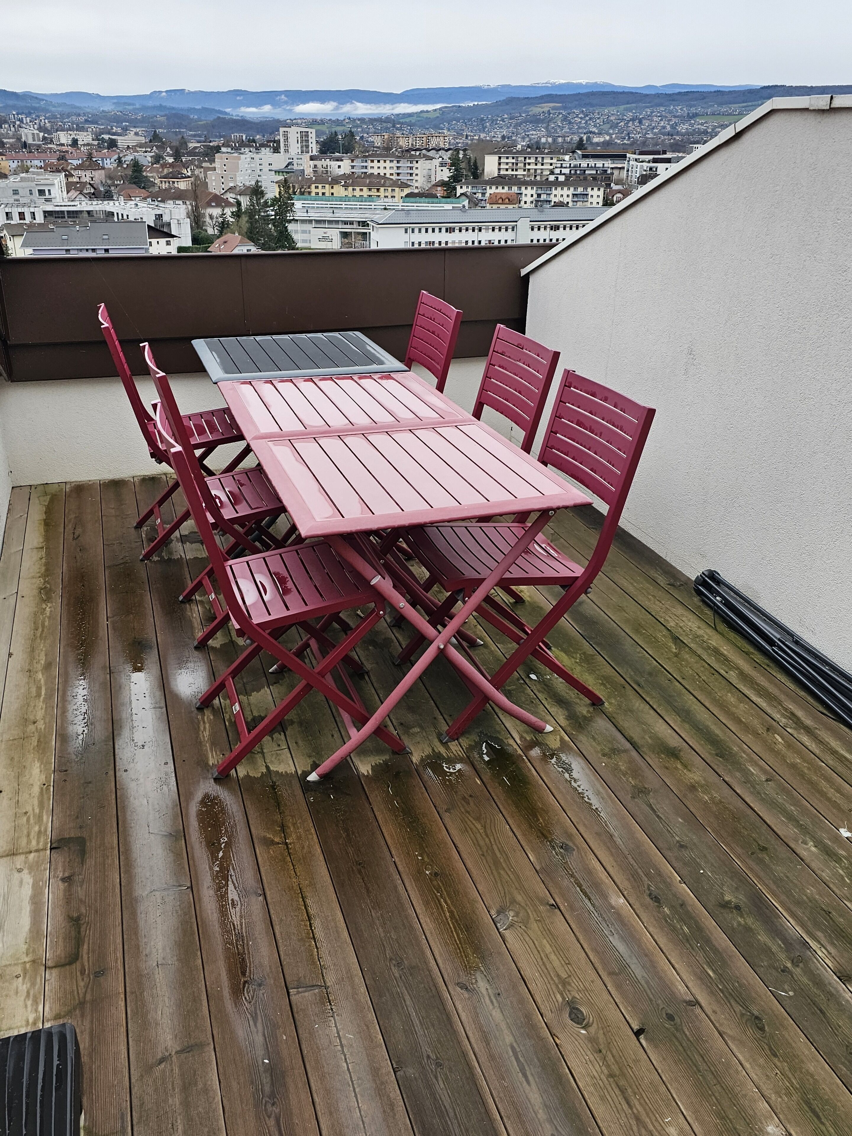Une table et des chaises en métal rouge, posées sur une terrasse en bois, avec une vue sur la ville en arrière-plan.
