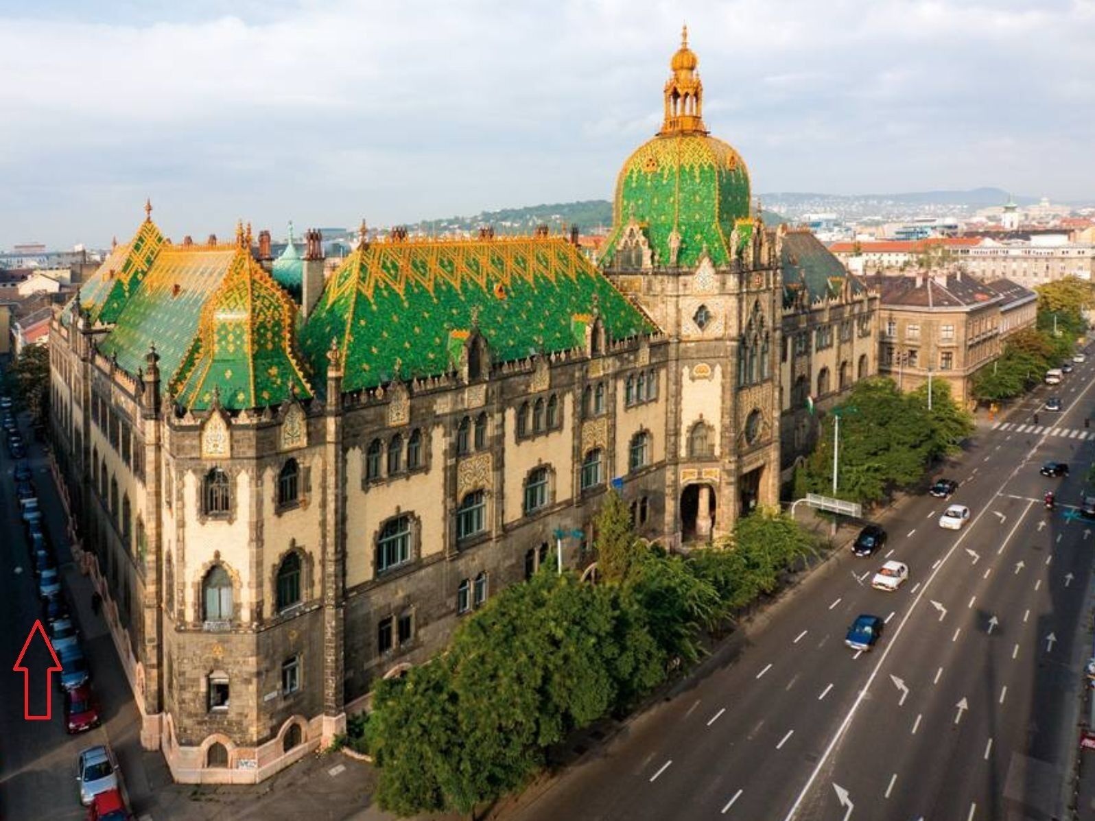 A large, historic building with a green dome and ornate roof, surrounded by a busy street and greenery.