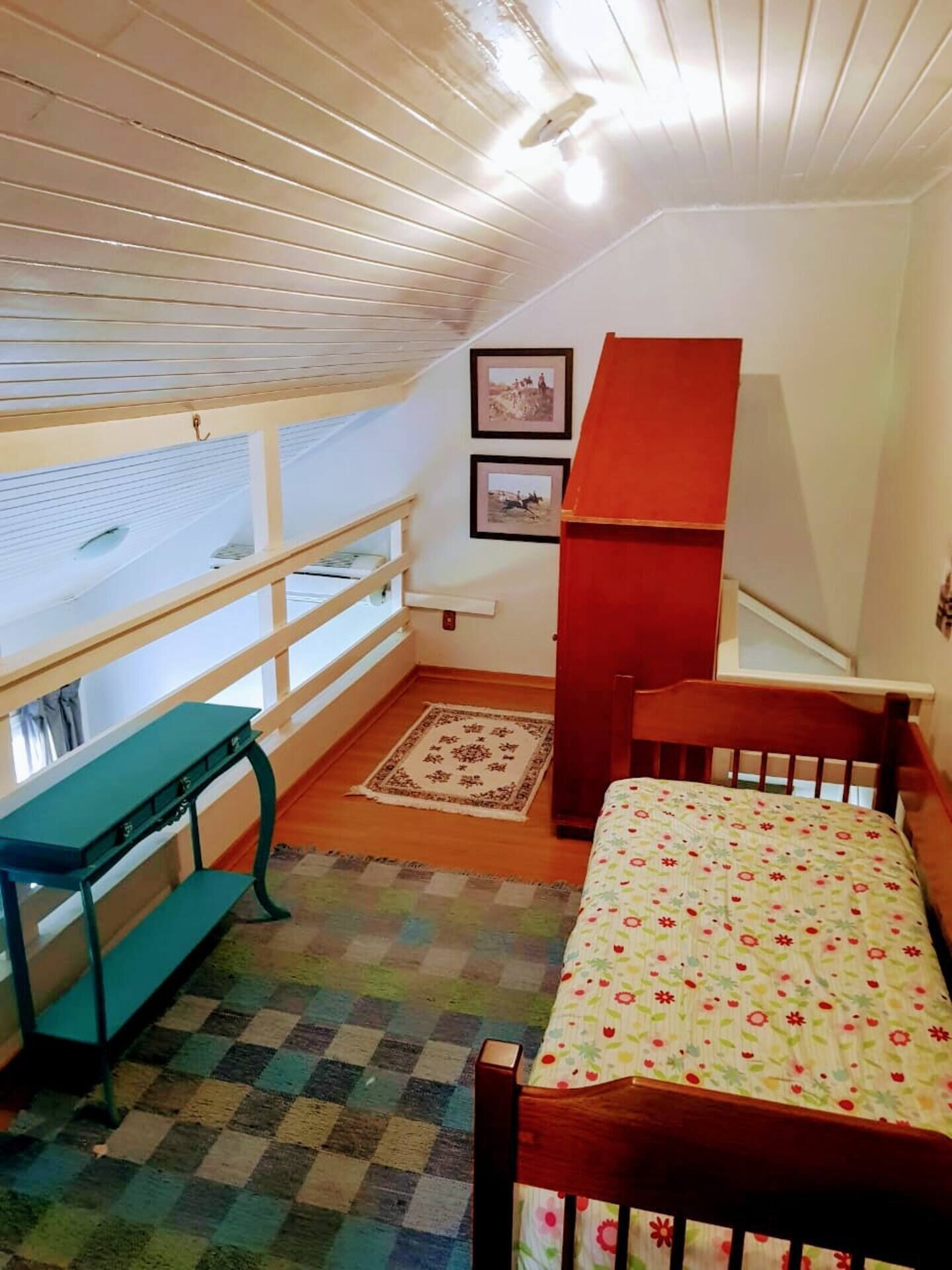 A small, cozy room with a wooden ceiling, a bed with a floral bedspread, a wooden bench, a small table, and a red cabinet.