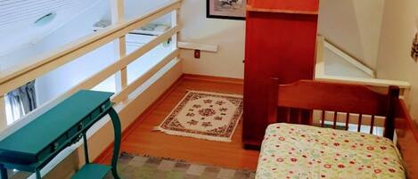 A small, cozy room with a wooden ceiling, a bed with a floral bedspread, a wooden bench, a small table, and a red cabinet.
