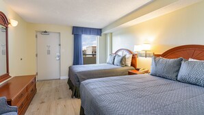 Ett hotellrum med två sängar, en byrå i trä, ett skrivbord med en lampa och ett fönster med utsikt över byggnader.