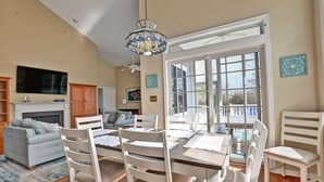 A dining area with a large table, white chairs, and a chandelier. There is a fireplace, a television, and a view of the outdoors through a large window.