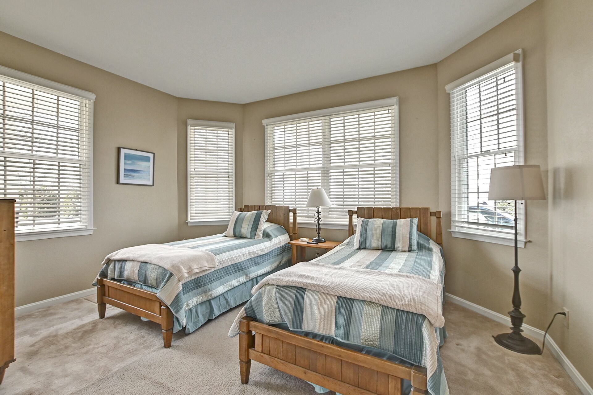 A bedroom with two wooden beds, beige walls, and large windows with blinds.