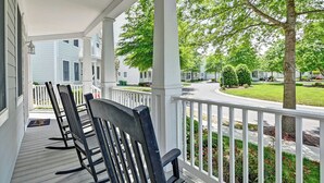 A covered porch with wooden chairs, white railings, and a view of a residential street with houses and trees.