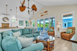 A spacious living room with a large sofa, a coffee table, and a dining area with chairs. There are ceiling fans, a view of the outdoors, and a large window. - Jersey Girl 5 Bedroom Home by RedAwning (Anna Maria)