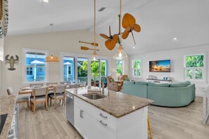 A spacious living area with a kitchen island, dining area, and a large window overlooking a blue building. - Jersey Girl 5 Bedroom Home by RedAwning (Anna Maria)