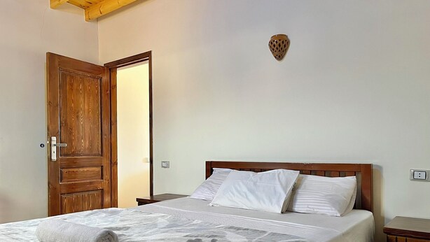 Een slaapkamer met een houten deur, een bed met wit beddengoed, een geborduurd plaid en een klein nachtkastje.