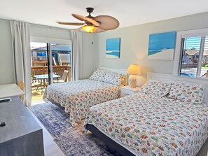 A hotel room with two beds, a desk, a ceiling fan, and a view of the outside.