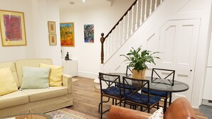 A living room with a sofa, a coffee table, a dining table with chairs, a staircase, and artwork on the walls. - Elegant 2 Bedroom Garden Maisonette near Olympia (London)
