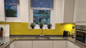 A modern kitchen with a sink, stove, and oven, white cabinets, a window with plants, and a yellow backsplash. - Elegant 2 Bedroom Garden Maisonette near Olympia (London)